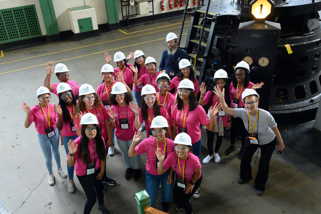 High school girls in STEM academy t-shirts and hard hats looking up at a photographer standing a floor above them